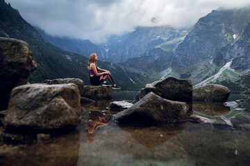 Young blonde fit and sporty woman in sportswear sitting on the rock on the shore and admiring beautiful view of green hills and mountains on Morskie Oko lake, High Tatras, Zakopane, Poland.