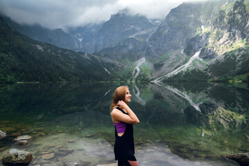 Beautiful young fit woman in sportswear standing on the rocky shore and admiring scenic view of green hills and mountains on Morskie Oko lake, High Tatras, Zakopane, Poland. © WellStock