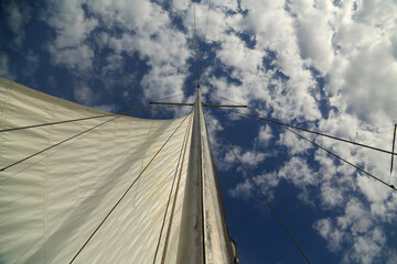 Fototapeta premium Mast of a yacht with a sail against the blue sky with white clouds. Walk on a yacht. Sailing holidays. Tourism. Travel by water. Regatta.
