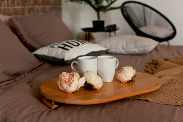 A wooden breakfast table in bed with two white cups and delicate cream peonies on a cozy bed of warm colors against a brick wall. Beautiful pillows with the inscription Home. Interior design. 