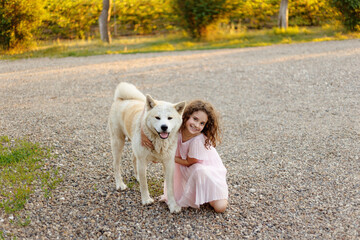 Cute little girl with a big white dog in the park. A beautiful 7 year old girl in pink dress hugs her favorite dog during a summer walk.