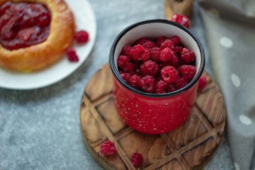 In the foreground is a red mug with garden raspberries, in the background a cheesecake with berry filling, light background, defocus; summer recipes, homemade vitamin snack