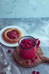 In the foreground is a red mug with garden raspberries, in the background a cheesecake with berry filling, light background, defocus; summer recipes, homemade vitamin snack