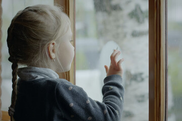Cute five year old girl draws on the window with fingers. Cute little girl drawing with her fingers on fogged window.