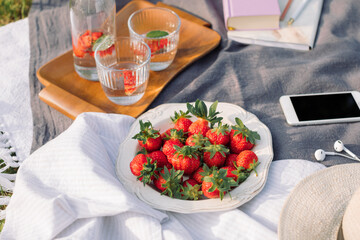 Summer picnic on the grass. Fresh strawberries,  refreshing drinks, mobile phone, headphones, hat, book on the blanket. Lifestyle concept.