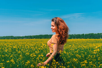 Naklejka na ściany i meble A woman with long hair walks in a rapeseed field, a photo taken from the back, the concept of flowering and summer day, a place for the text banner