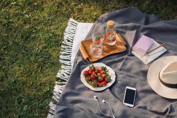 Summer picnic on the grass. Book, fresh strawberries, refreshing drinks, mobile phone, headphones, hat on the blanket. Lifestyle concept.