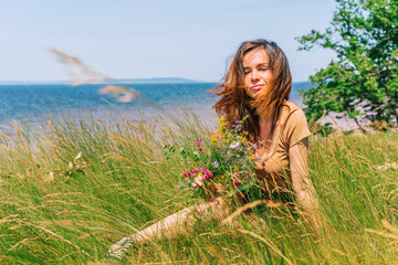 A happy woman with long hair sits in the grass and holds a bouquet of wild flowers against the blue sky