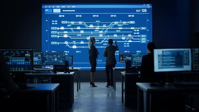 Project Manager and Computer Science Engineer Talking while Using Big Screen Display Showing Infrastructure Infographics and Data.Telecommunications Company System Control and Monitoring Room