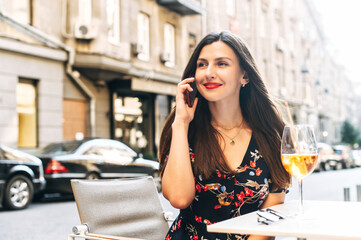 Fototapeta premium Smiling happy woman is talking on the phone while sitting in the city cafe and drinking a cocktail, she looks away