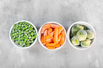 Frozen vegetables such as green peas, brussels sprouts and baby carrot in the white bowls on the concrete gray background