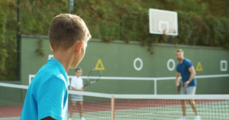 Rear of Caucasian teen boy playing big tennis with joyful handsome father and small sister outdoors in summer. Back view on teenage play with dad and girl at sport court. Family weekend.