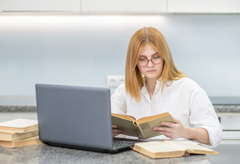 Fototapeta premium Smart teen girl read a book and uses laptop at home. Distance learning concept