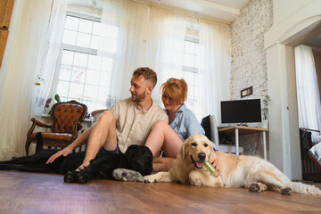 young couple with dogs sitting on the floor of their flat and having fun. dogs playing with their toys. man and woman hug each other and spend leisure  time with their dogs