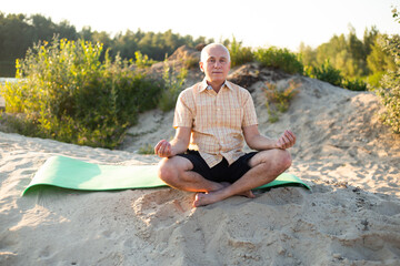 Healthy senior man meditating in beach, sitting lotus pose on yoga mat, zen