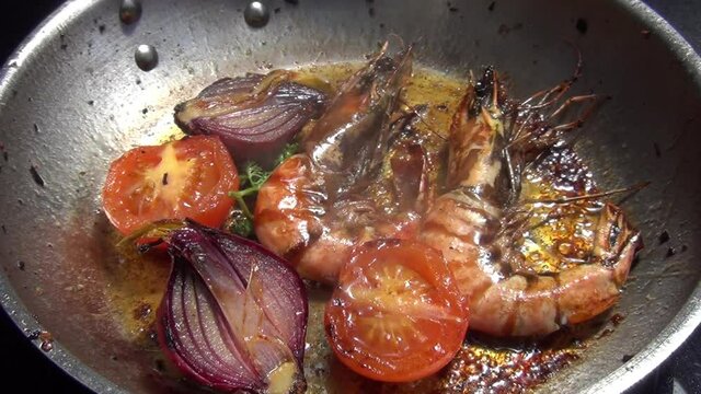 Shot of two tiger prawns, red onion tomatoes and curly parsley which are cooking in melted garlic butter in a stainless-steel pan