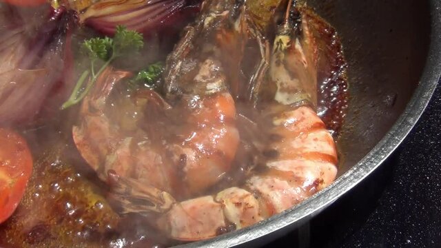 A zoom in footage of two tiger prawns, red onions, tomatoes and curly parsley sizzling in melted garlic butter in a stainless-steel pan