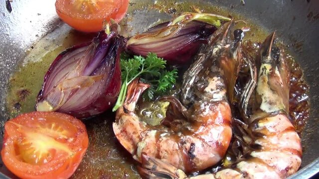 Slow zoom out shot of two tiger prawns, red onion tomatoes and curly parsley cooking in melted garlic butter in a stainless-steel pan