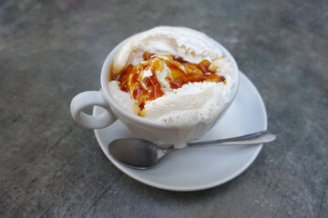 Close up white cup of cappuccino with delicious foam covered with caramel topping on grey wooden table