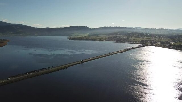 Drone footage of cars driving along long road to bridge surrounded by blue river on sunny winter day with low mountains in background in Bridgewater, Tasmania, Australia.