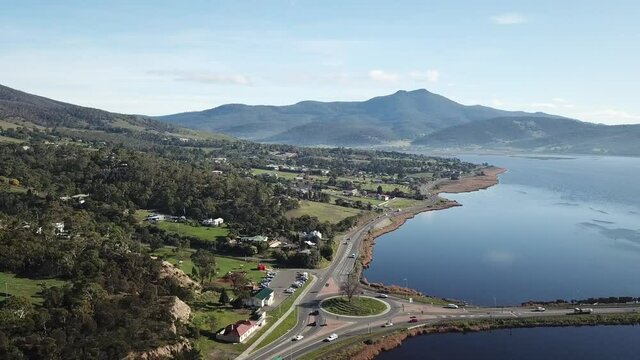 Slow drone pan of cars driving through round about surrounded by blue river on sunny winter day with low mountains in background in Hobart, Tasmania, Australia.