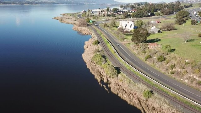 Drone footage of bike and car driving down solo road along small coast town in Hobart with clear blue water on bright sunny winter day, Tasmania, Australia.