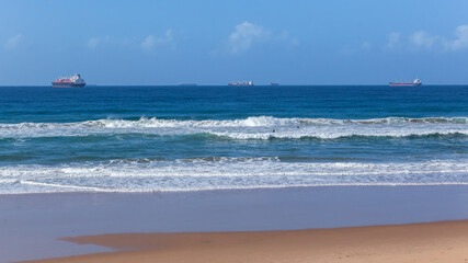 Fototapeta na wymiar Beach Ocean Waves Crashing Breaking With Cargo Ships Anchored on Sea Horizon.