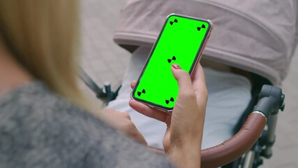 A young mother is rocking a baby stroller with one hand and flipping through a green-screen phone with the other. A girl flips through a smartphone with a chromakey on the background of a stroller