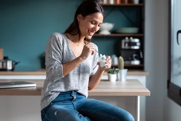 Fotobehang Smiling young woman eating yogurt while sitting on stool in the kitchen at home. © nenetus
