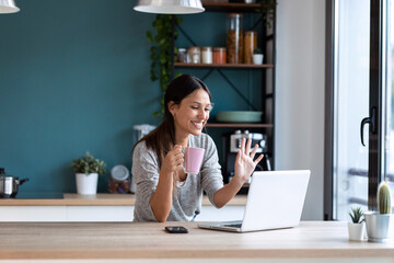 Smiling young woman waving through the laptop web camera while holding a cup of coffee in the kitchen at home.