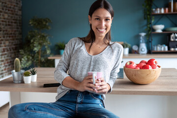 Smiling young woman looking at camera while drinking a cup of coffee in the kitchen at home.