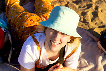 A pretty girl in a hat looks up at the camera and smiles, lying on her stomach on a Mat on the beach on the sand. She is dressed in a yellow jumpsuit, a white t-shirt and a blue Panama hat