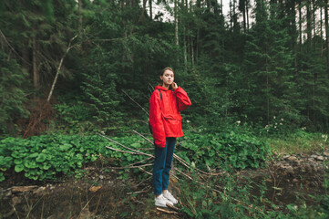 Tourist girl standing on a tree trunk over the mountain river, looking at the camera, feeling calm and relaxed in the woods. Young female touching her hair, enjoying the peaceful nature of the forest.