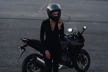 Obraz premium Biker woman in black leather jacket and full face helmet sits near stylish sports motorcycle at urban parking. Traveling and active lifestyle concept.