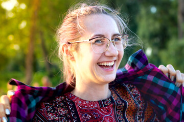 A young beautiful cute happy girl with long hair and glasses laughs sincerely, a big wide smile on her face. She's wearing a plaid shirt. In nature against the background of green trees. Outdoor