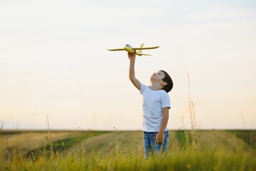 Happy child playing with a toy plane in nature during summer sunset. Boy in a white shirt with a plane in hands on wheat field. Kid holds a wooden airplane and dreams of being a pilot, on the nature