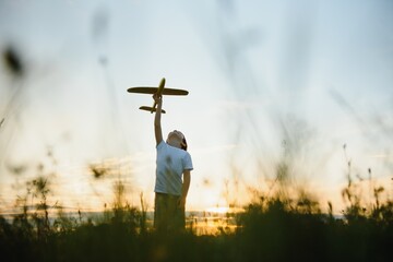 Happy child playing with a toy plane in nature during summer sunset. Boy in a white shirt with a plane in hands on wheat field. Kid holds a wooden airplane and dreams of being a pilot, on the nature