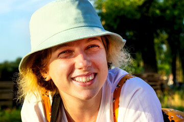 Close-up of a cute hipster girl sincerely smiling, laughing, looking at the camera. She's wearing a white t-shirt and a blue Panama hat. Summer outdoor recreation. Emotion of joy and happiness