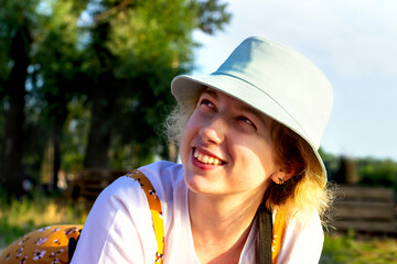 Close-up of a cute hipster girl sincerely smiling, laughing, looking away. She's wearing a white t-shirt and a blue Panama hat. Summer outdoor recreation. Emotion of joy and happiness
