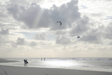 Schattenhaftes Paar und Kitesurfer am Strand im Gegenlicht - die Sonne scheint durch die Wolkenschicht auf das silbernglänzende Meerwasser