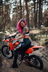 Obraz na płótnie Canvas Beautiful young hipster girl with pink hair and tattoo on hand in sports bra and leather pants, smiling and looks down, sitting on a motorcycle in off road adventure