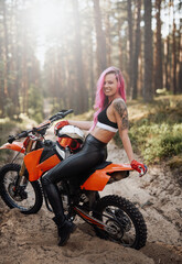Fototapeta na wymiar Beautiful young hipster girl with pink hair and tattoo on hand sitting on her modern motocross bike in woods, smiling and looking on the camera, illuminated by bright sunlight
