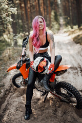 Fototapeta na wymiar Happy young woman with pink hair and tattoo on hand sitting on her modern motocross bike in woods