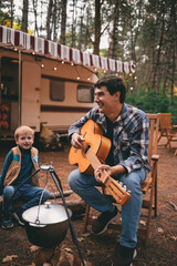 Happy family on a camping trip relaxing in the autumn forest. Camper trailer. Young man plays guitar by the fire