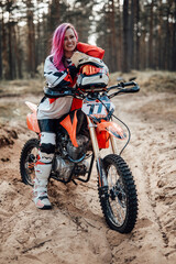 Naklejka premium Portrait of a smiling young female racer with pink hair in motocross kit sitting on her motorcycle in off road adventure