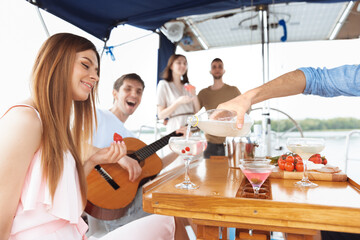 Group of happy friends drinking vodka cocktails at boat party outdoor. Young people playing guitar in sea tour, youth and summer vacation concept. Alcohol, vacation, resting. Focus on pouring drink.