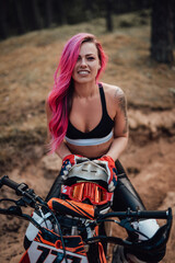 Fototapeta na wymiar Portrait of a charming young female racer with pink hair and tattoo on hand, smiling and looking on the camera while sitting on modern motorcycle in off road adventure