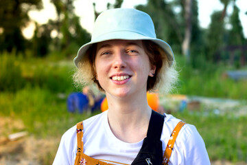A cute funny hipster girl looks thoughtfully at the camera, dressed in a white t-shirt and a blue Panama hat. In nature against the background of green trees. Summer holidays and vacations. Happiness