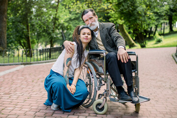 Young social worker or granddaughter takes care about senior man in wheelchair at walk in a park, smiling and embracing each other, looking at camera. Different generations of people