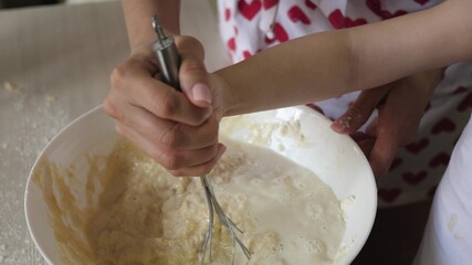 Mom teaches a child to cook dough. Mother and kid beat with a whisk a mass of flour with milk. Cooking at home with family. Teamwork